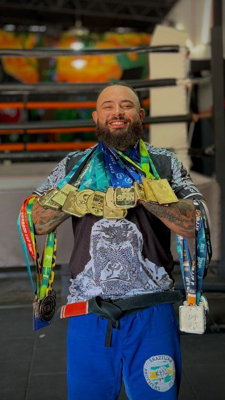 Uriel Meneses de faixa preta, sorrindo, carregando uma pilha de medalhas no pescoço e nos braços