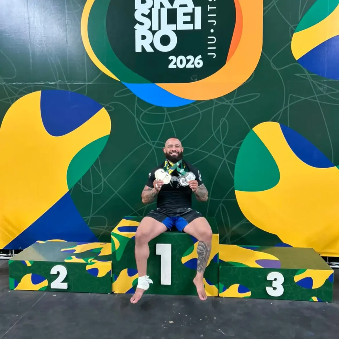 Uriel Meneses sentado no lugar mais alto do pódio do Campeonato Brasileiro 2026, com duas medalhas e o pé direito enfaixado