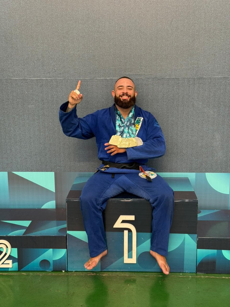 Uriel Meneses de kimono azul sentado no lugar mais alto do pódio, com medalhas no pescoço e o dedo indicador para cima